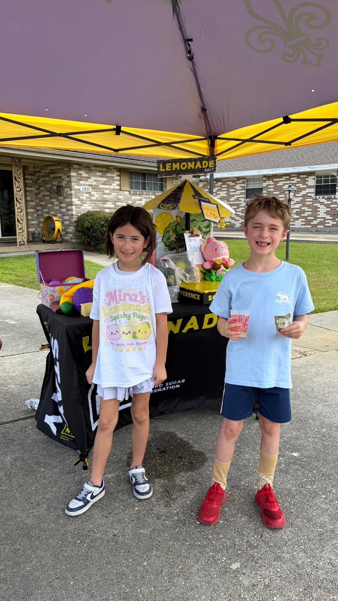 Mira and her best friend standing in front of the yellow Mira's Lemonade Squishy tent, smiling with a cup of fresh-squeezed lemonade and a dollar bill from a happy customer