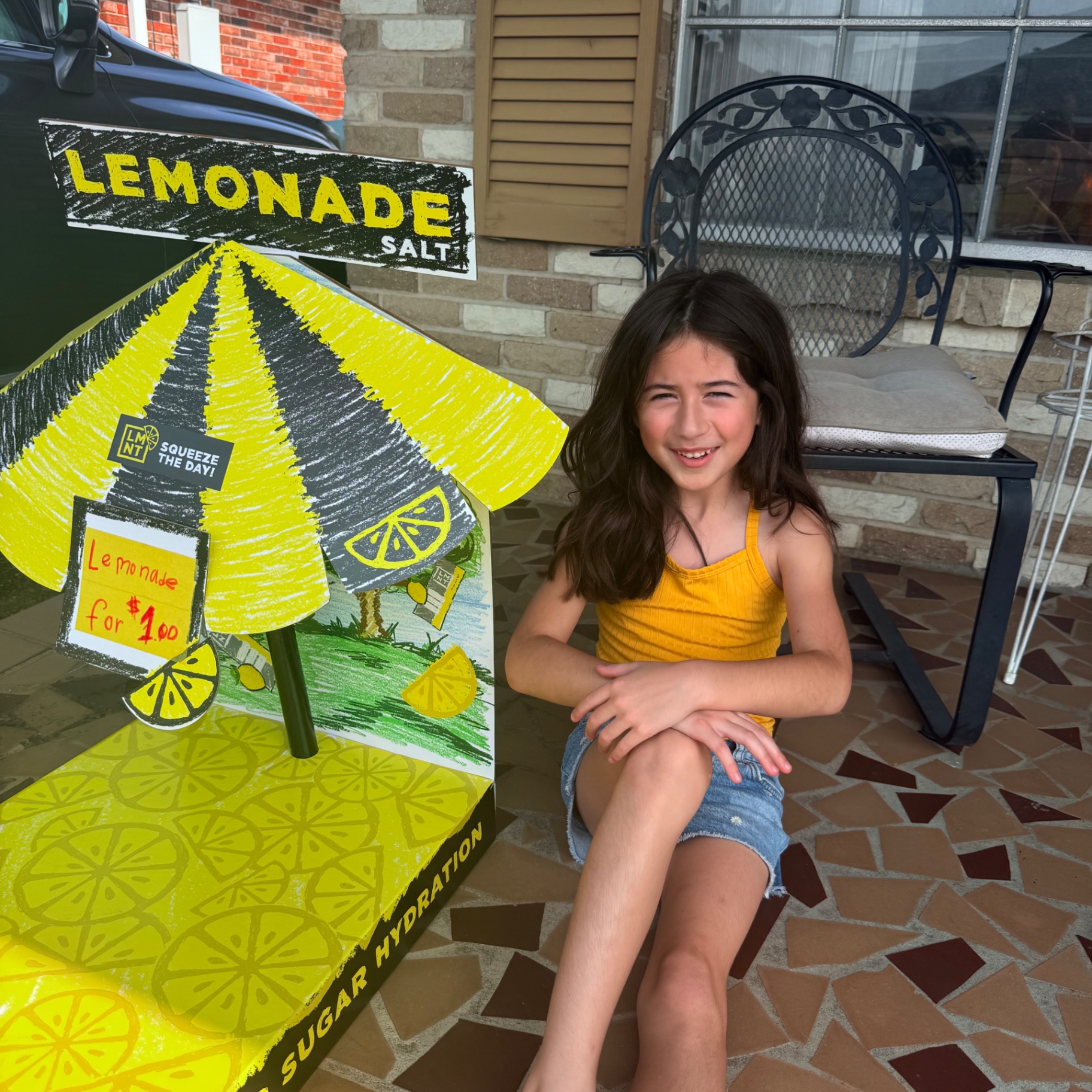 Young kid entrepreneur Mira smiling proudly next to her homemade yellow cardboard lemonade stand sign reading Lemonade $1.00 on the front porch — the very first Mira's Lemonade Squishy pop-up stand