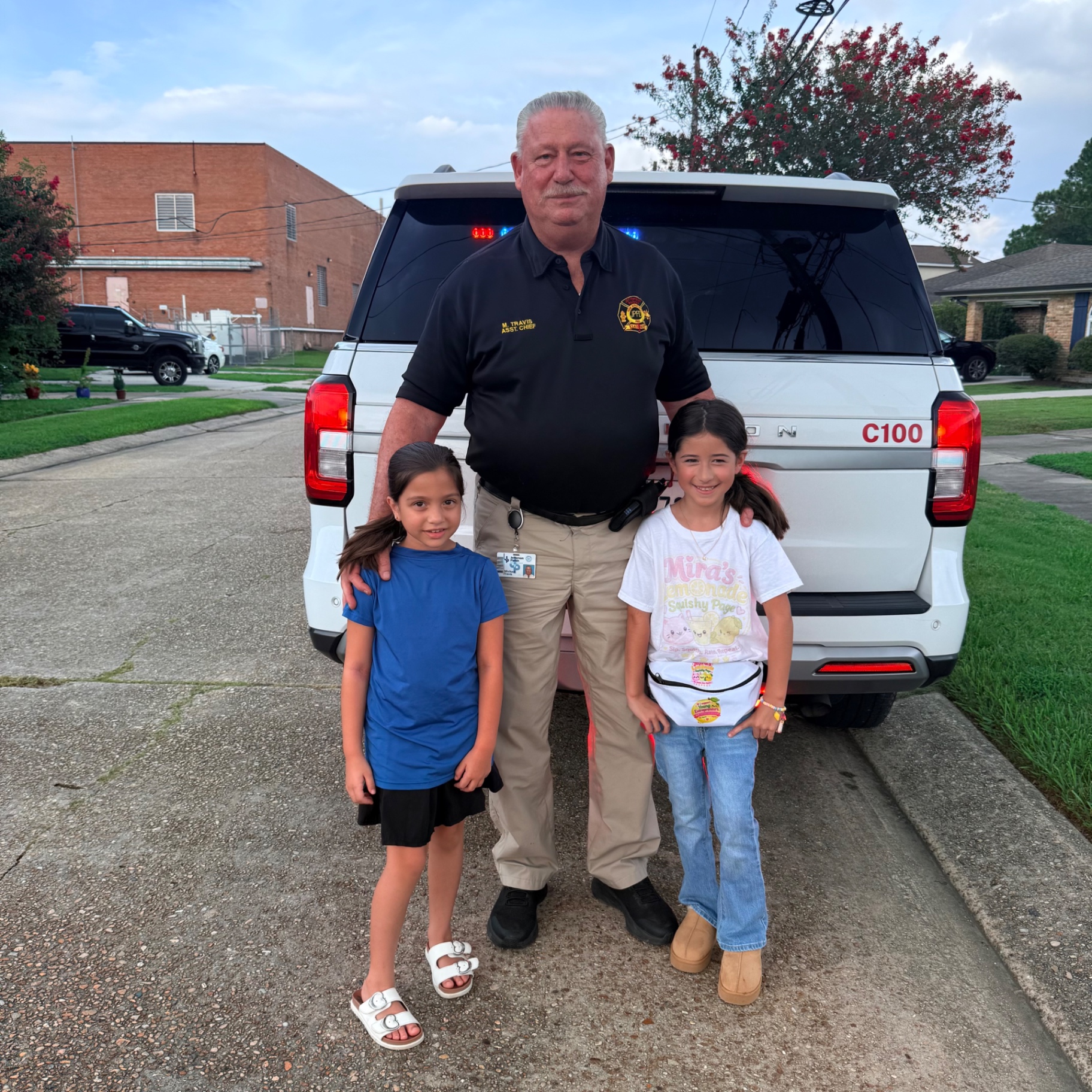 Jefferson Parish Fire Department Assistant Chief M. Travis standing with Mira in her Mira's Lemonade Squishy shirt and squishy fanny pack and her cousin in front of the white C100 command truck in Metairie, Louisiana