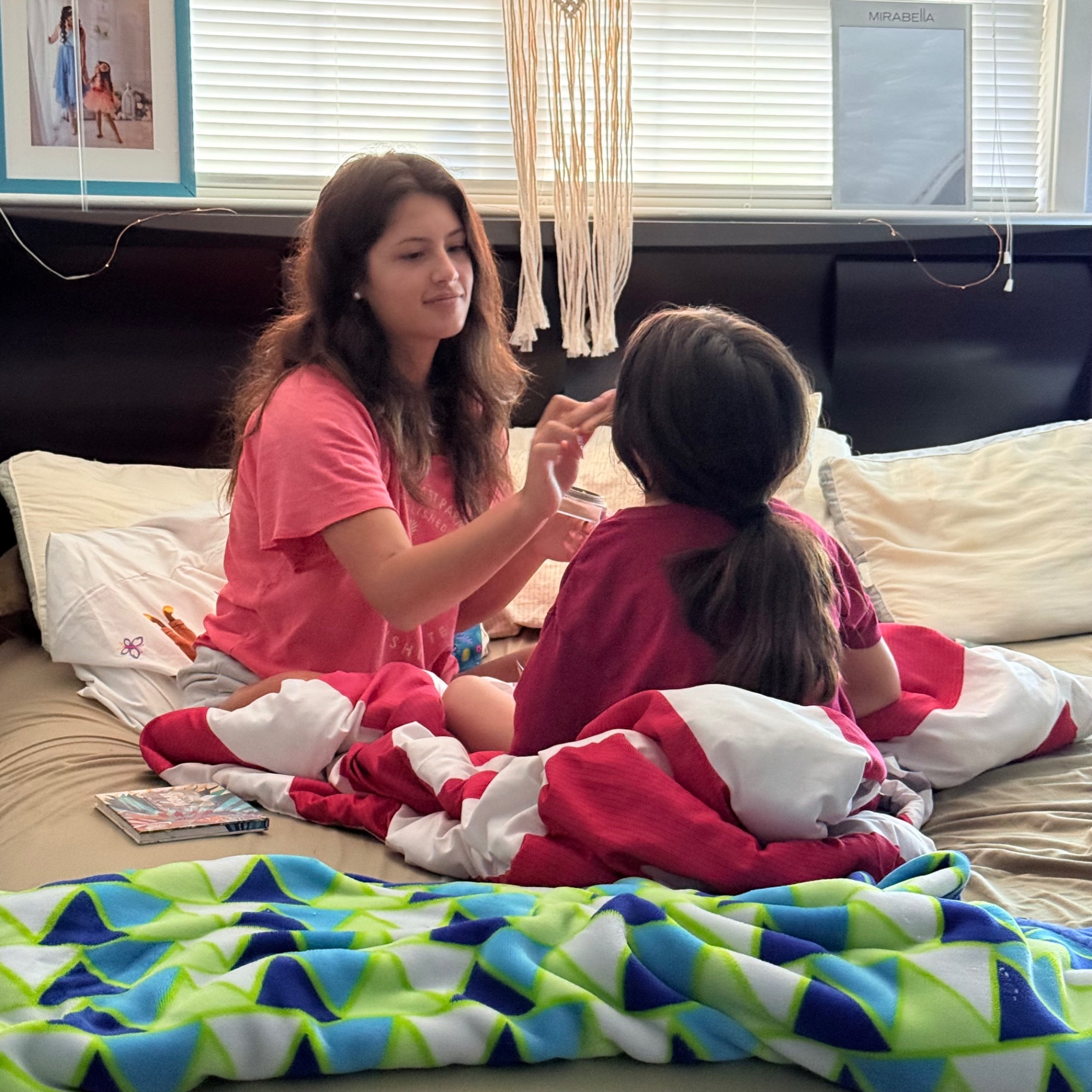 Mira in a pink t-shirt sitting on the bed doing a playful skincare and dress-up session with her little sister in a red and white outfit, gently applying cream to her face during a cozy summer sisters day at home