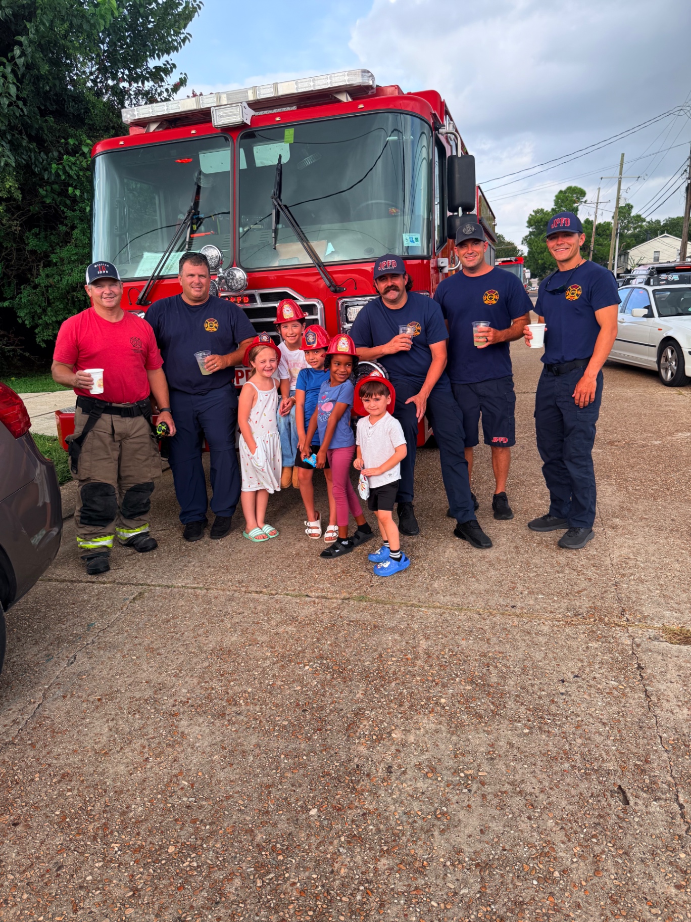 Five Jefferson Parish firefighters holding cups of lemonade beside a red JPFD fire engine while six kids in red plastic fire helmets, including Mira in her Mira's Lemonade Squishy shirt, crowd in front for a group photo at the Metairie lemonade stand and squishy rating table