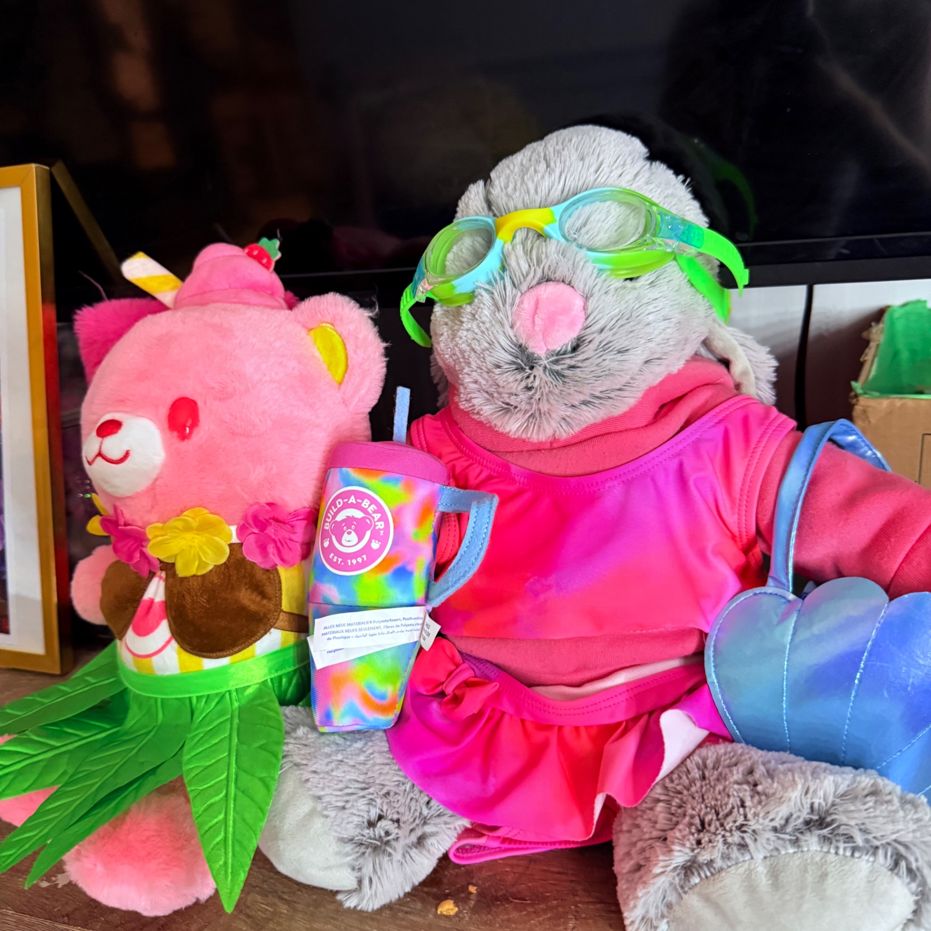 A soft pink teddy bear plushie sitting beside a gray bunny plushie wearing pink swim goggles, a pink dress, and holding a tie-dye Build-A-Bear cup — part of Mira's plushie and squishy collection