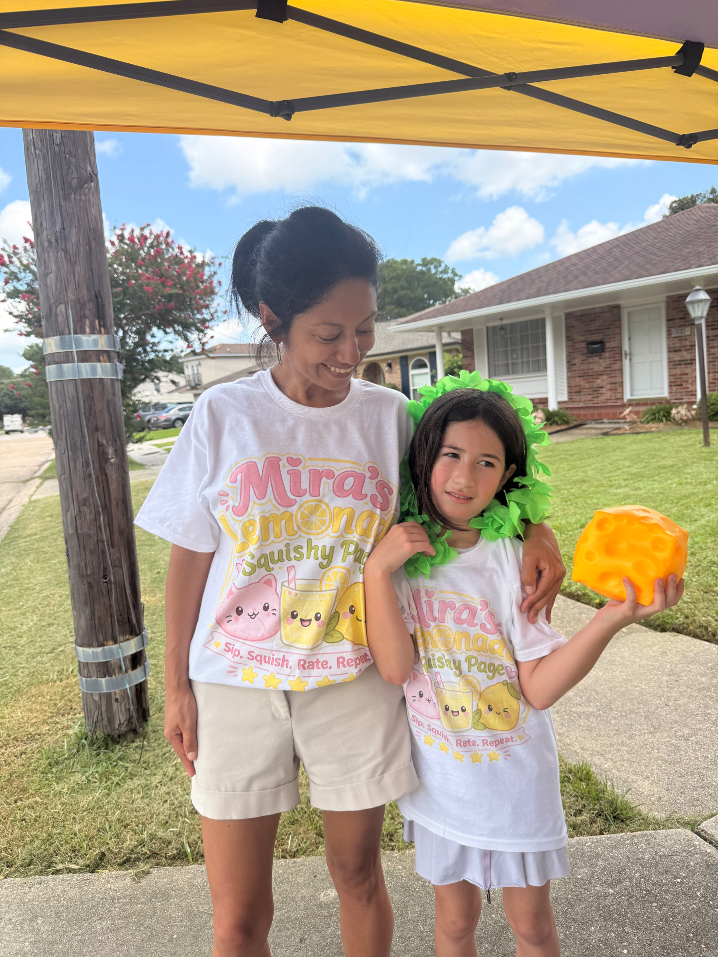Mira and her mom under the yellow Mira's Lemonade Squishy tent in matching brand t-shirts, Mira hugging a giant orange cheese-wedge squishy while running the stand together